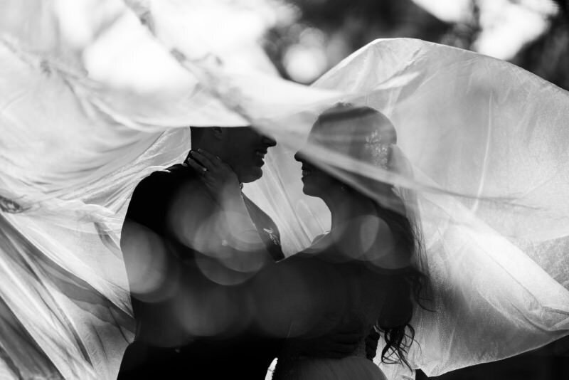 a black and white image of the wind catching a brides veil as she dances with her groom on their wedding day