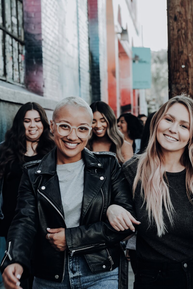 Four women of different nationalities walking together smiling with one women in the front has her arm through the other woman's arm next to her.
