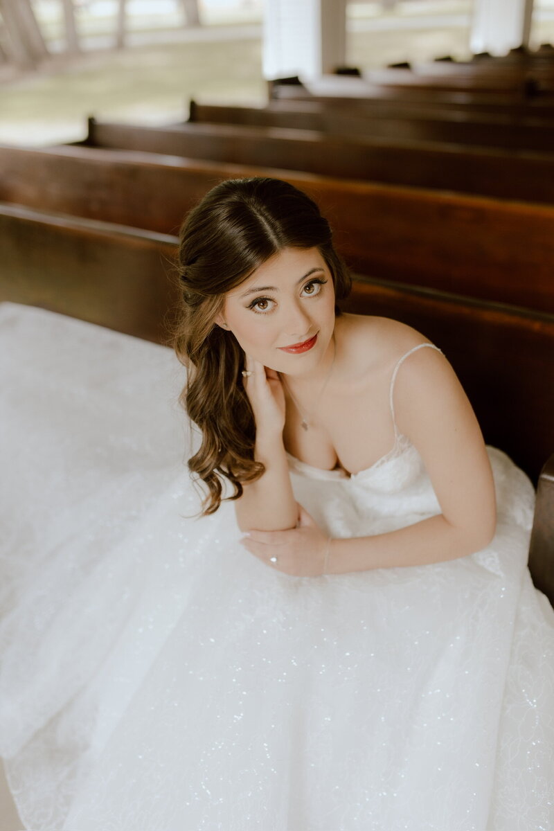Bride in a white gown sitting on a wooden pew, looking up with a soft smile, as her bridal hair and makeup showcase the artistry of exclusive Texas wedding packages.