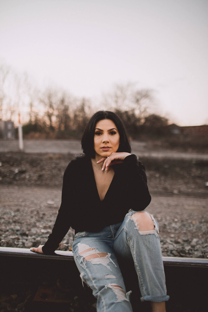Angelica Ray, a confident woman in ripped jeans and a black top, sitting outdoors at dusk, symbolizing empowerment and resilience on a spiritual journey.