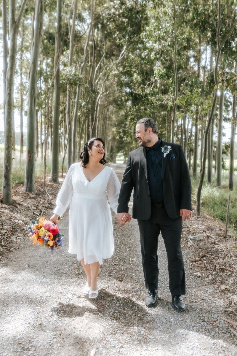 A bride and groom walking down a road lined with trees and holding a bright bouquet