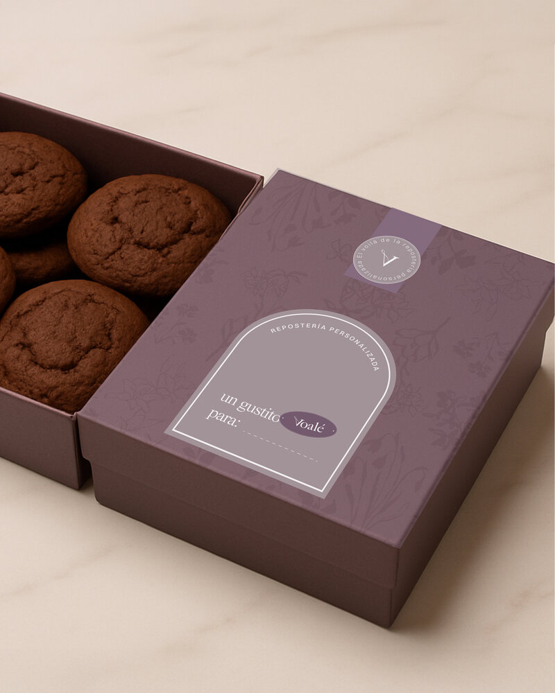 Close-up of a refined purple gift box from Voalé Bakery, partially opened to reveal artisanal chocolate cookies. The lid features delicate floral patterns and a personalized label. The branding conveys a sense of warmth, intention, and elevated craftsmanship.
