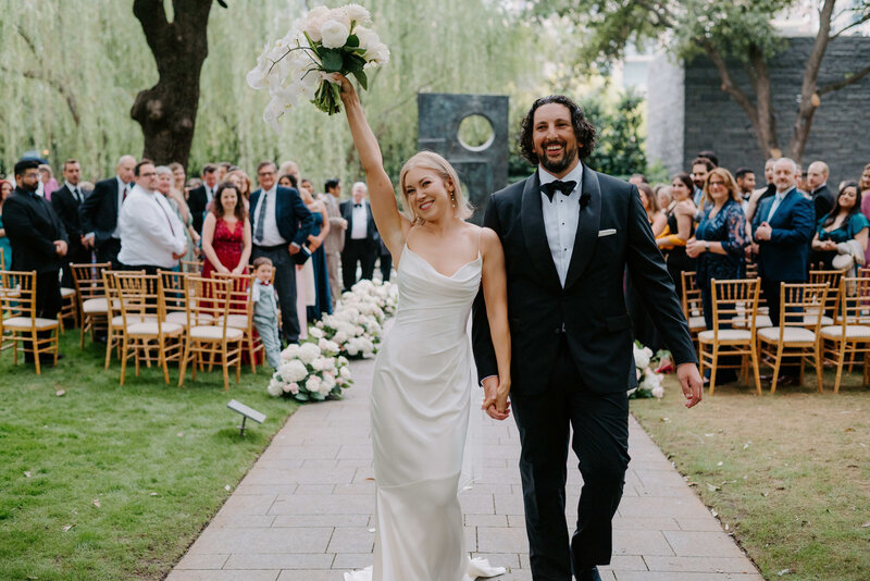 Bride and groom walk joyfully down the aisle after outdoor wedding ceremony, surrounded by cheering guests