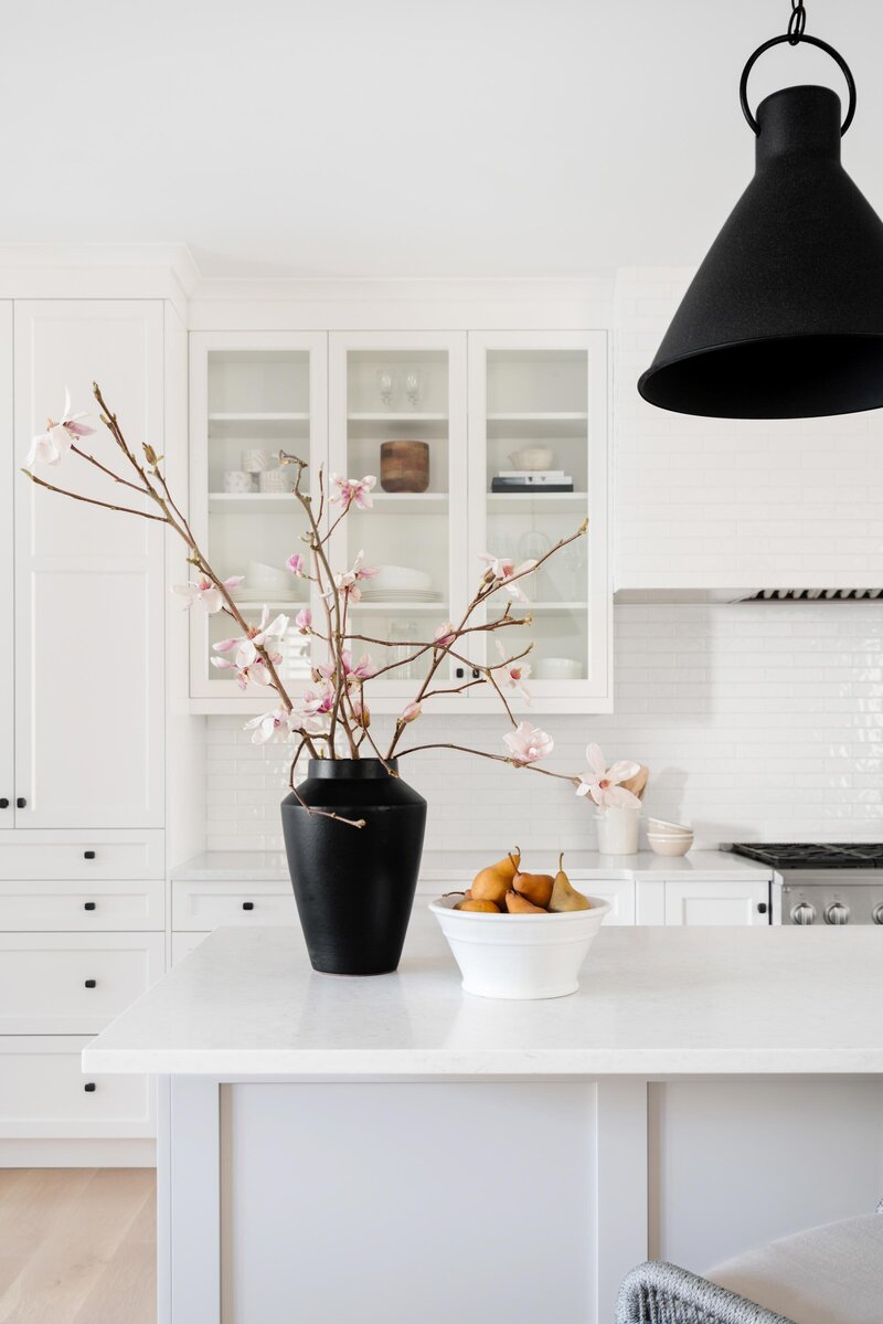 white kitchen cabinets, white kitchen island and countertops, facing range, large black vase with cherry blossom branches, large black pendant light, white bowl with golden pears