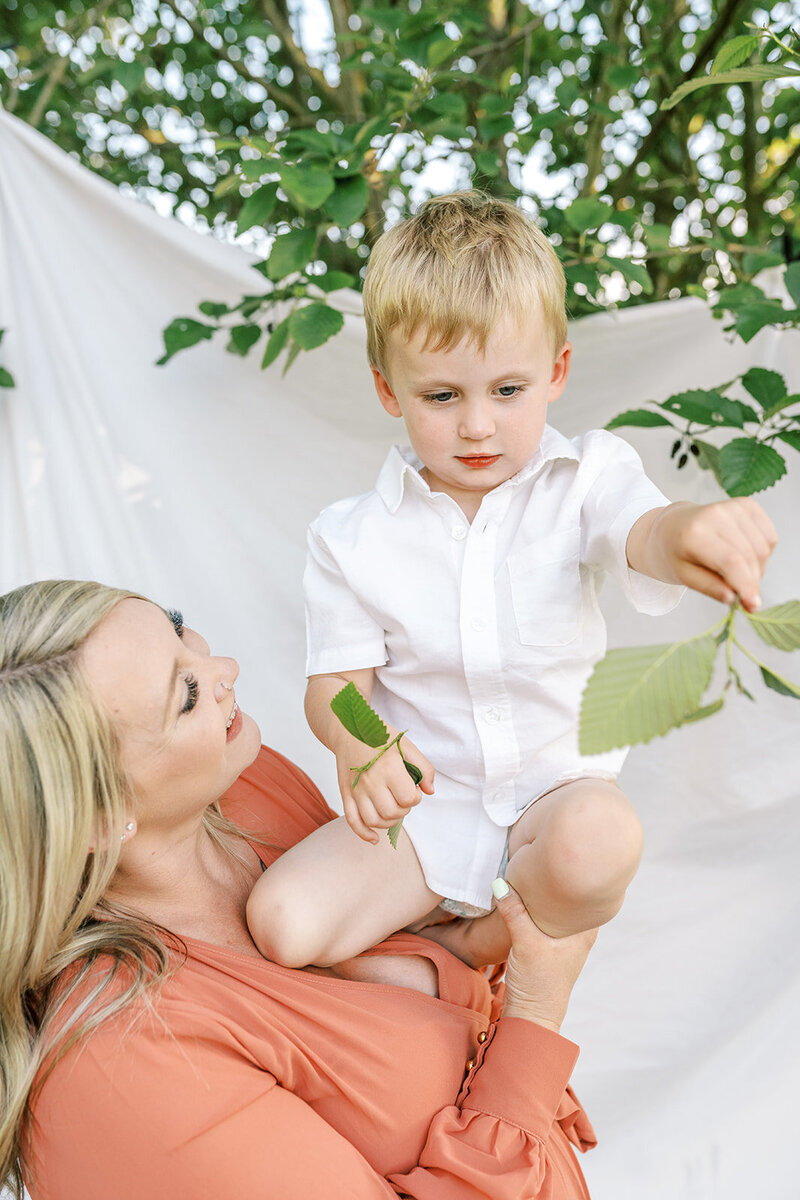 A mom holds a toddler up in her arms, representing the family photography portraits style of Among the Oaks. 