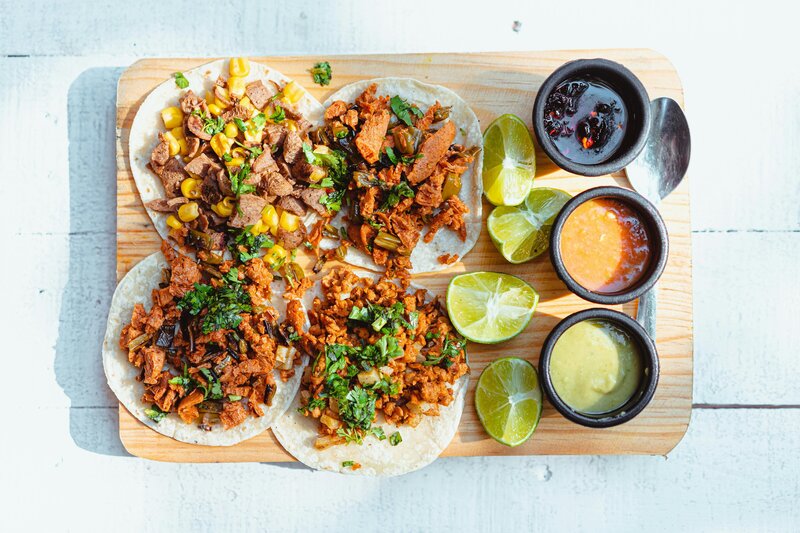 A wooden platter holds four tacos filled with assorted toppings including meats, vegetables, and herbs. Lime wedges are placed around the tacos. On the right side, there are small bowls containing various sauces, including red, green, and dark sauces.
