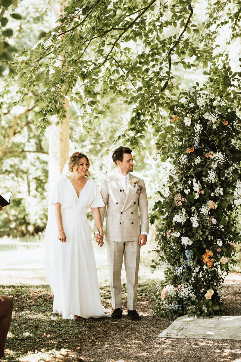 Bride and groom standing by a broken arch of flowers at the outdoor ceremony area at Broadfield Court