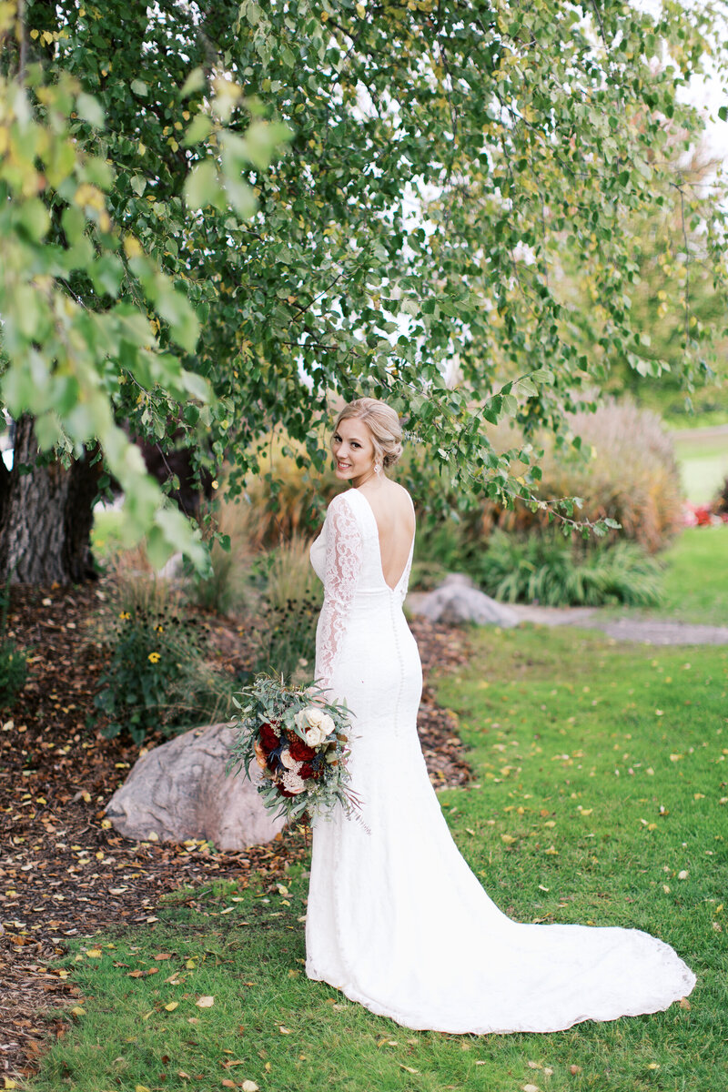 Bride with her gorgeous bouquet  looking over her shoulder with a smile under the tree. Captured by Toly Dzyuba, Minnesota wedding photographer. 