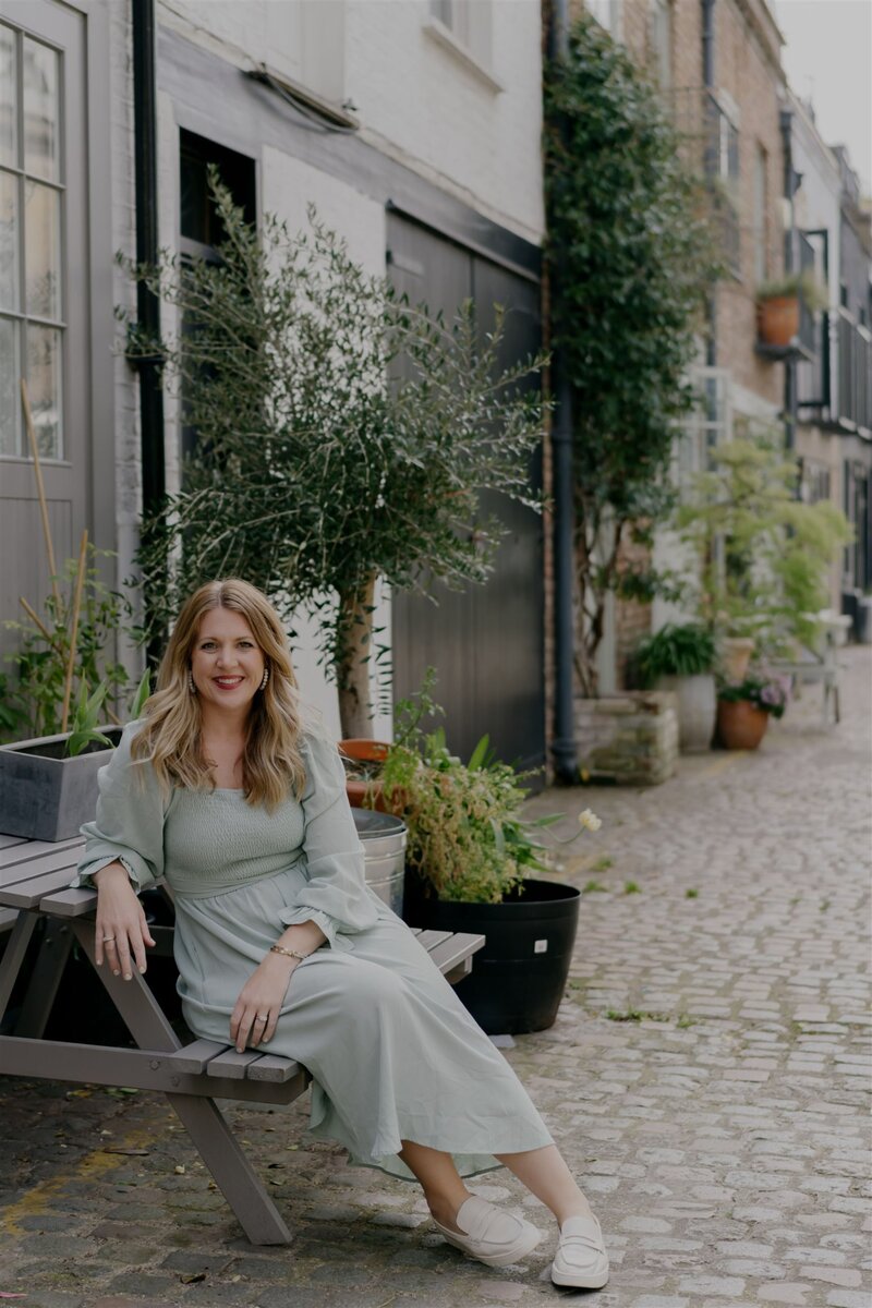 Dana smiling in a light green dress sitting on a bench in a charming European-style alley lined with potted plants and rustic buildings.
