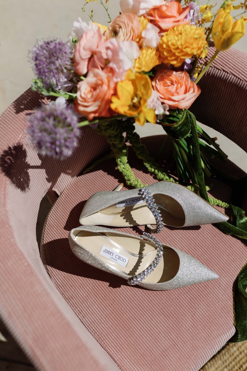 Jimmy Choo wedding shoes styled on pink velvet chair with colorful bouquet.