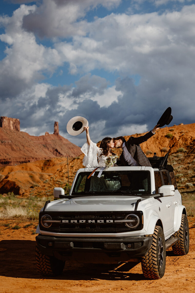 A couple hangs out the sunroof of their Bronco on their wedding day in Moab