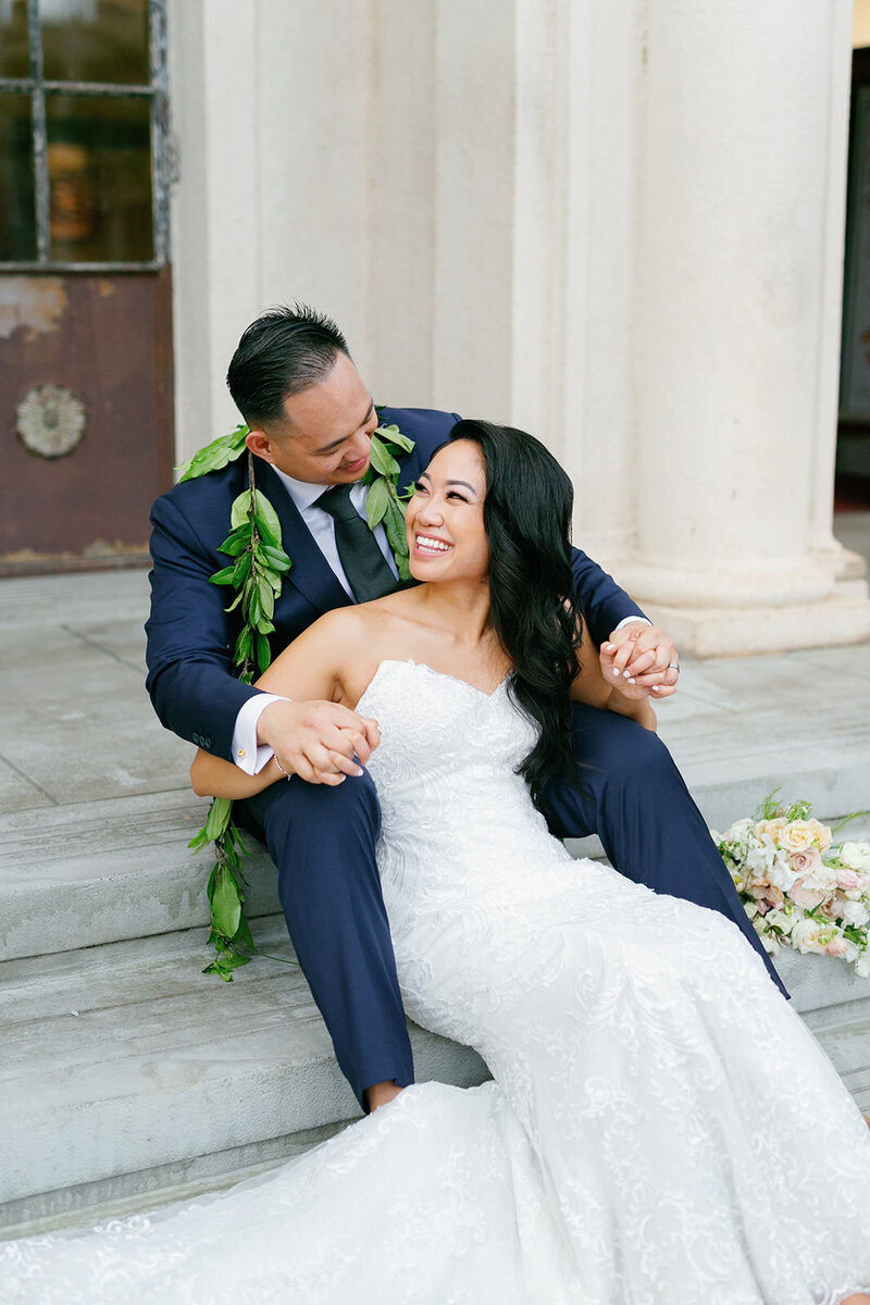 A newlywed couple sitting on a staircase one in front of the other smiling at each other 