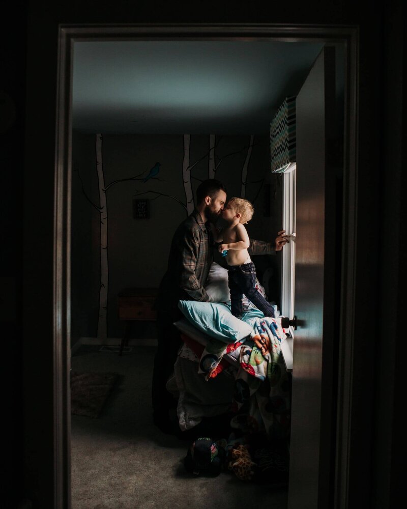 Toddler kisses his dad  while standing on the bed next to a window. Shot by family photographer Tracy Miller.