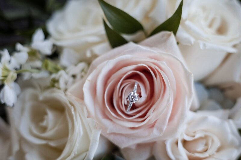 Close-up of a sparkling diamond engagement ring nestled inside a blush rose, surrounded by ivory roses and greenery at a Jewish wedding at JPS Events in downtown Indianapolis.