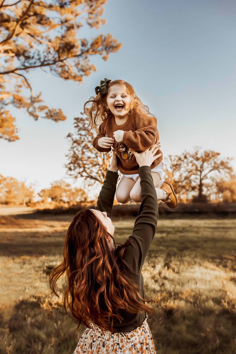 A woman lifts a laughing child into the air in a grassy field with autumn trees in the background captured by a family photographer in Macon GA.