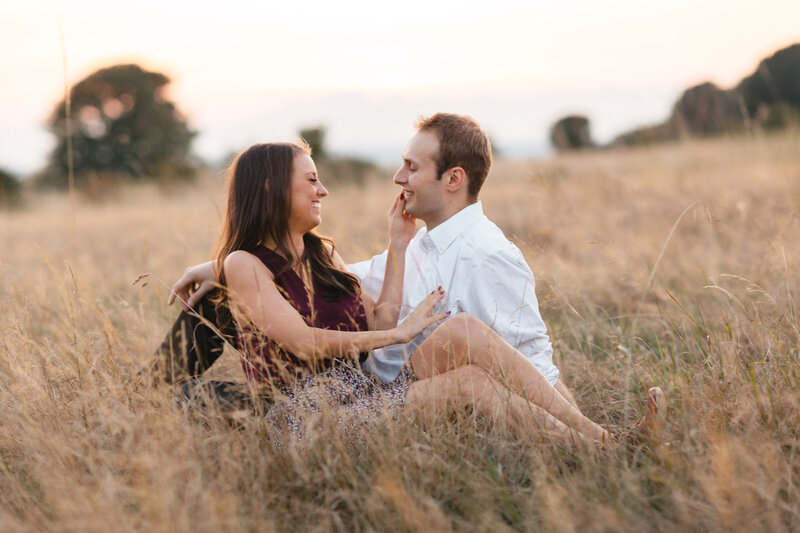 Engaged couple sitting in grassy fields of Discovery Park in Seattle at sunset.