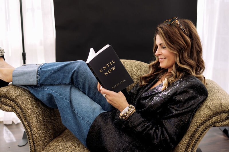Kerri Link sits comfortably in a chair, smiling as she reads her book Until Now.