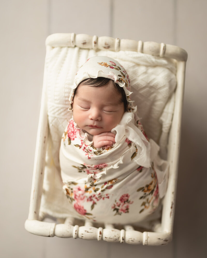 Newborn girl styled in full floral wrap and bonnet, posed on a soft cream bed in vintage tones.