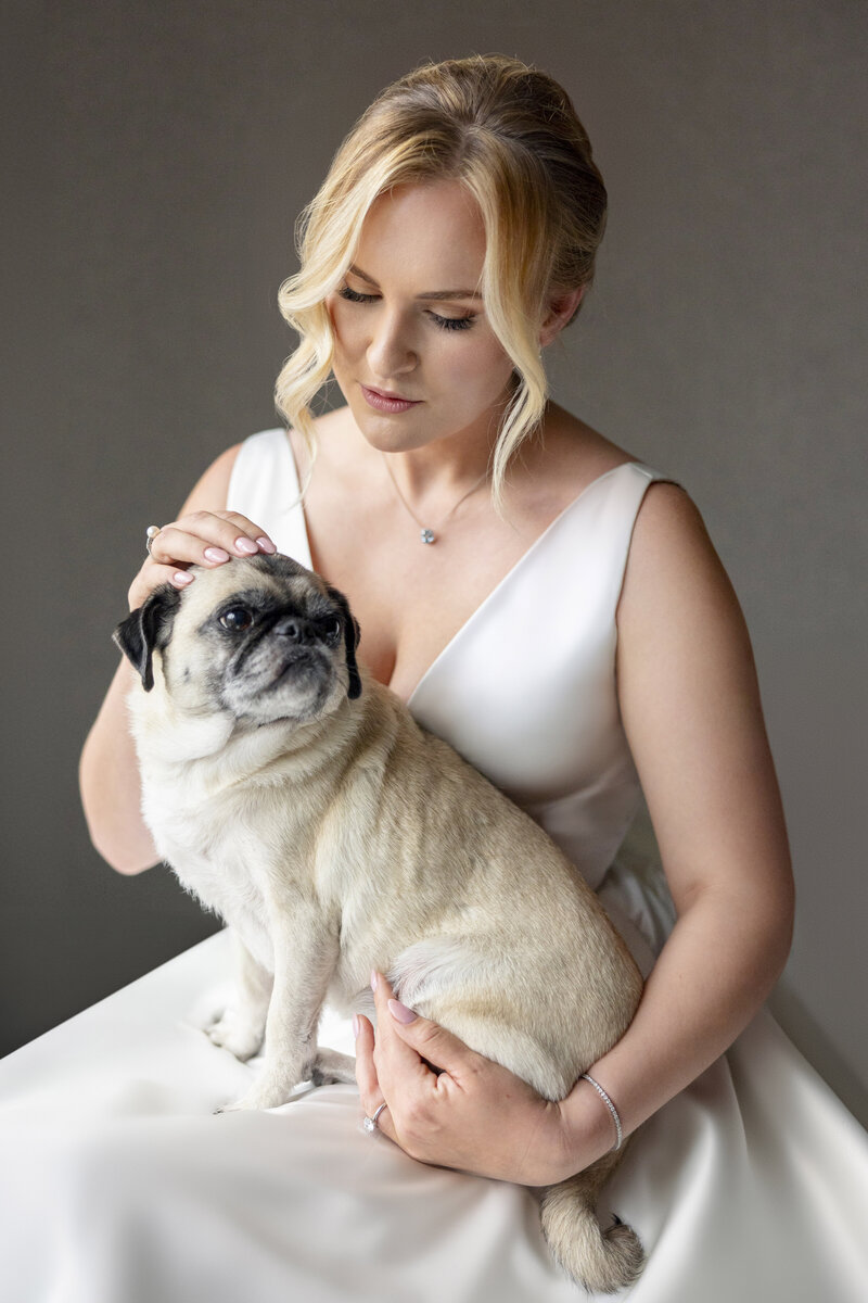 A woman in a white dress gently holds and pets a pug on her lap. The scene is serene and the background is neutral, highlighting the tender moment between the woman and the dog.