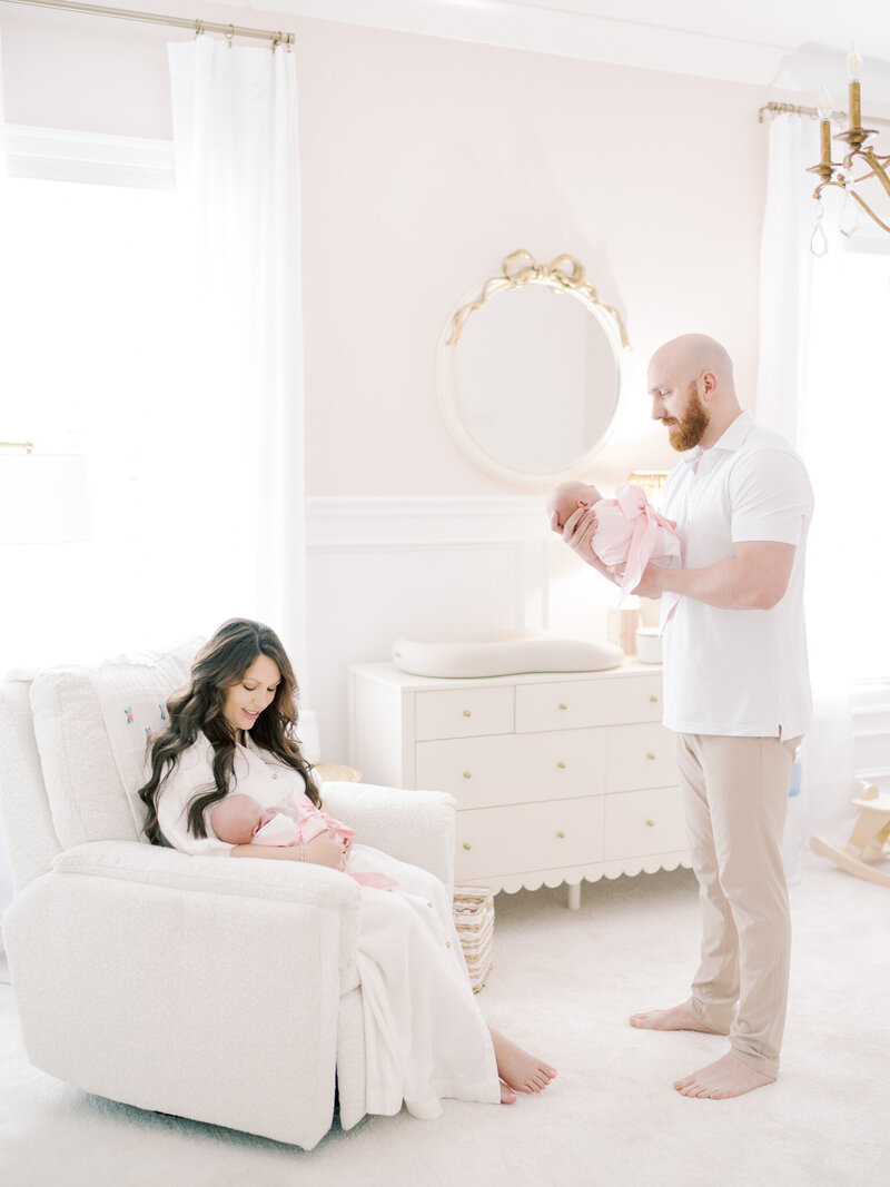 Brunette motMother in rocking chair and father standing in nursery each holding a twin newborn girl wrapped in swaddlesher in cream, floral dress lying on couch reaches down to touch her swaddled, newborn son sitting in a moses basket in the floor of their Little Rock home taken by Little Rock newborn photographer Bailey Feeler Photography