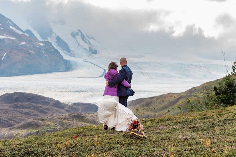 A couple in formal wedding attire stand in an embrace on the green top of a mountain.  Dress train, coat tails, and hair blow in the stiff wind.  They look out to a massive glacier that stretches below them in a deep mountain valley as far as the eye can see