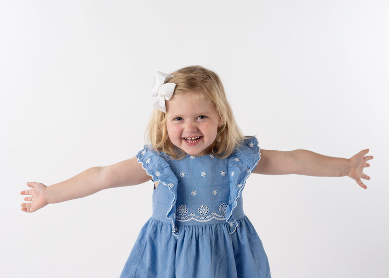 A blonde preschool age girl smiling for his school portrait in a blue dress.