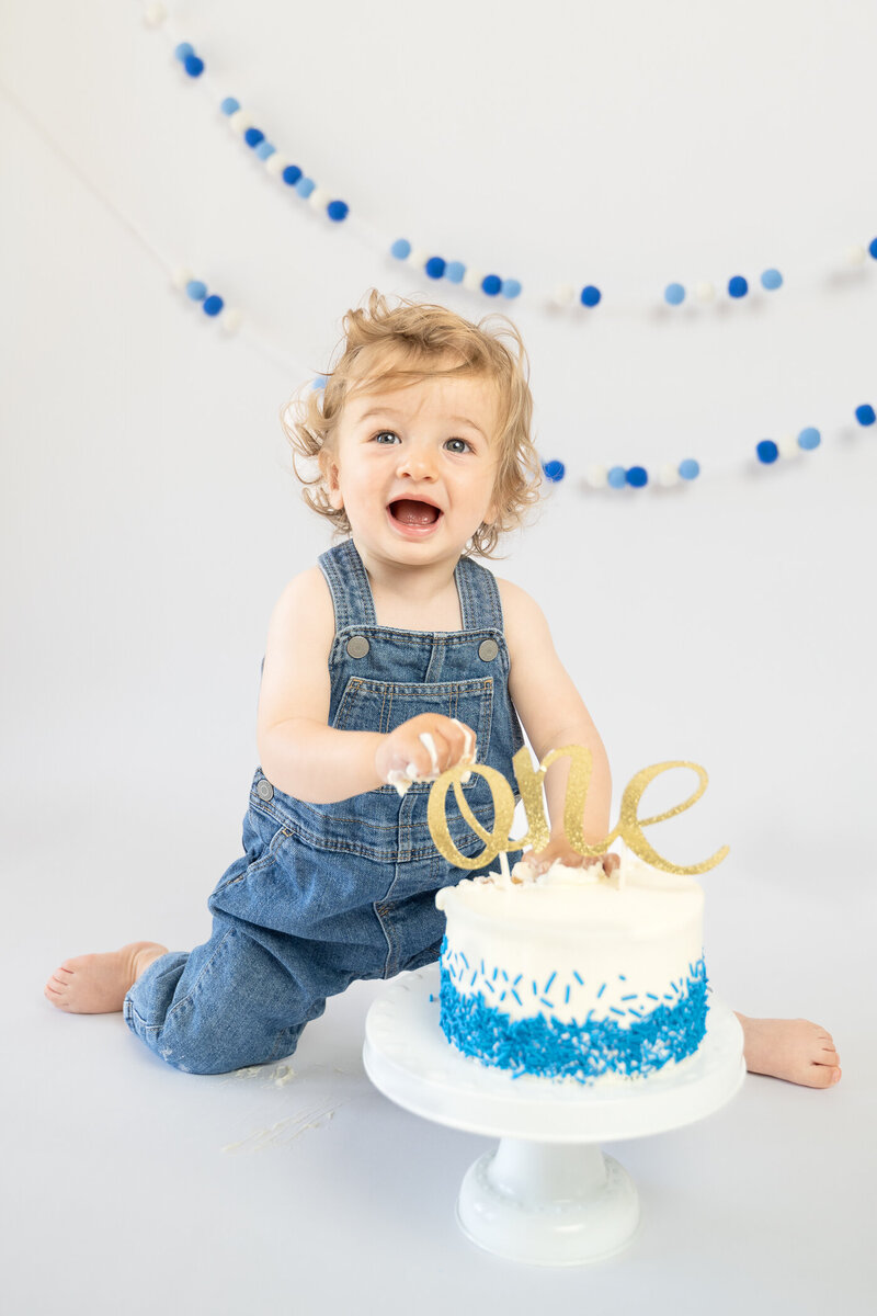 One year old boy in overalls touching a cake.