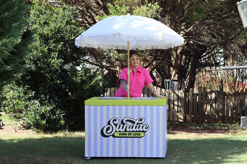 A woman in a pink jumpsuit standing behind a striped ice-cream cart.