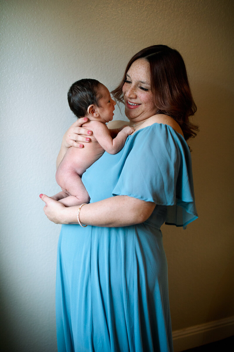 A woman stands in front of a window, holding her brand new baby, and wearing a lovely blue dress