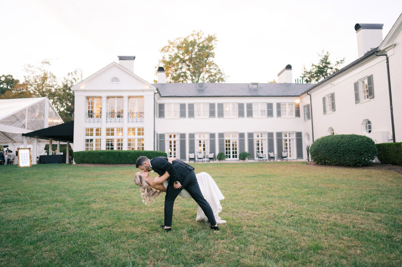 Maddie and Garrett pose for their wedding photos on the lush green lawn of Boxwood Estate. Maddie wears a stunning white wedding gown with intricate lace details, while Garrett is dressed in a sharp suit. The couple stands together, smiling and holding hands, surrounded by the estate's beautiful landscaping and tall trees. The soft, natural light adds a romantic touch to the scene, capturing a joyful and intimate moment between the newlyweds.