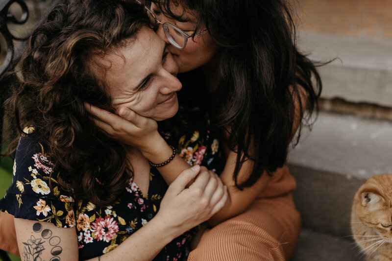 One woman kisses the cheek of the other while holding her face tenderly. They are seated on porch steps with a ginger cat nearby, both women displaying affection and warmth.