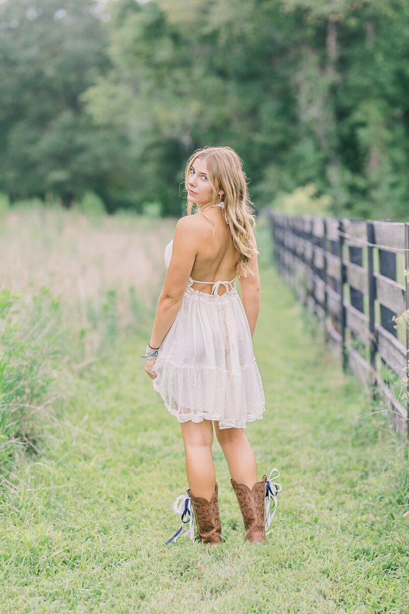 Outdoor senior portrait of a girl in a cream dress and cowboy boots walking along a wooden fence, captured by Raleigh Senior Photographer Lindsey Lambert Photography.