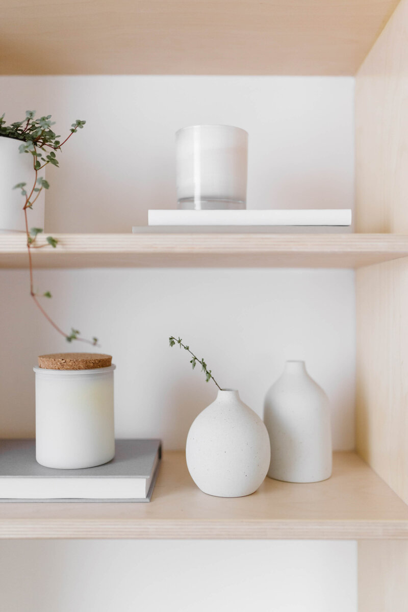 close-up view of plywood bookshelf, small white candle and book, green plant in white vase, two small organic-shaped white vases, gray book