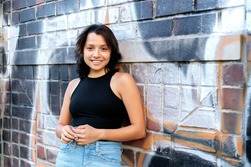 Senior girl in black sleeveless top and blue jeans leaning against graffiti wall outside during sunset