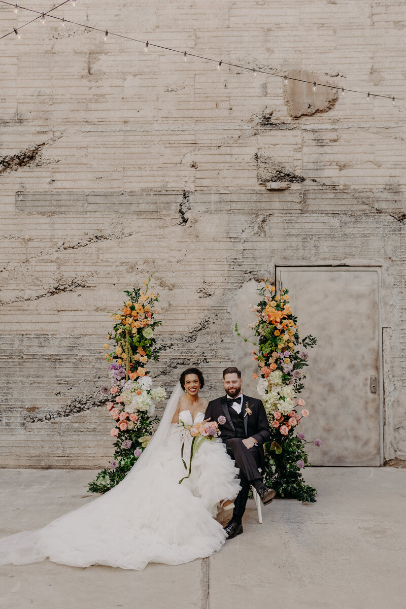 bride and groom sit side by side between floral arch at the ice house