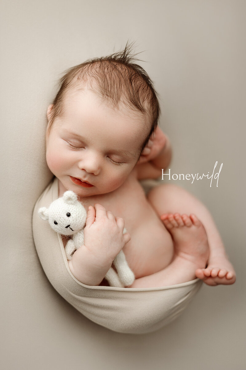 Wrapped newborn baby smiling sweetly, wearing a hand-knit cream hat and holding a stuffed animal. Photographed in a warm and cozy Grand Rapids, MI, studio for luxury newborn portraits