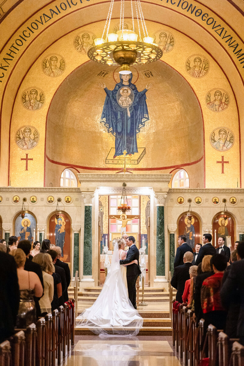 A couple shares a first kiss in a church during their wedding ceremony. The church features ornate gold and red decor, religious iconography, and an archway with an illuminated chandelier above. Guests are seated and standing in the aisles.