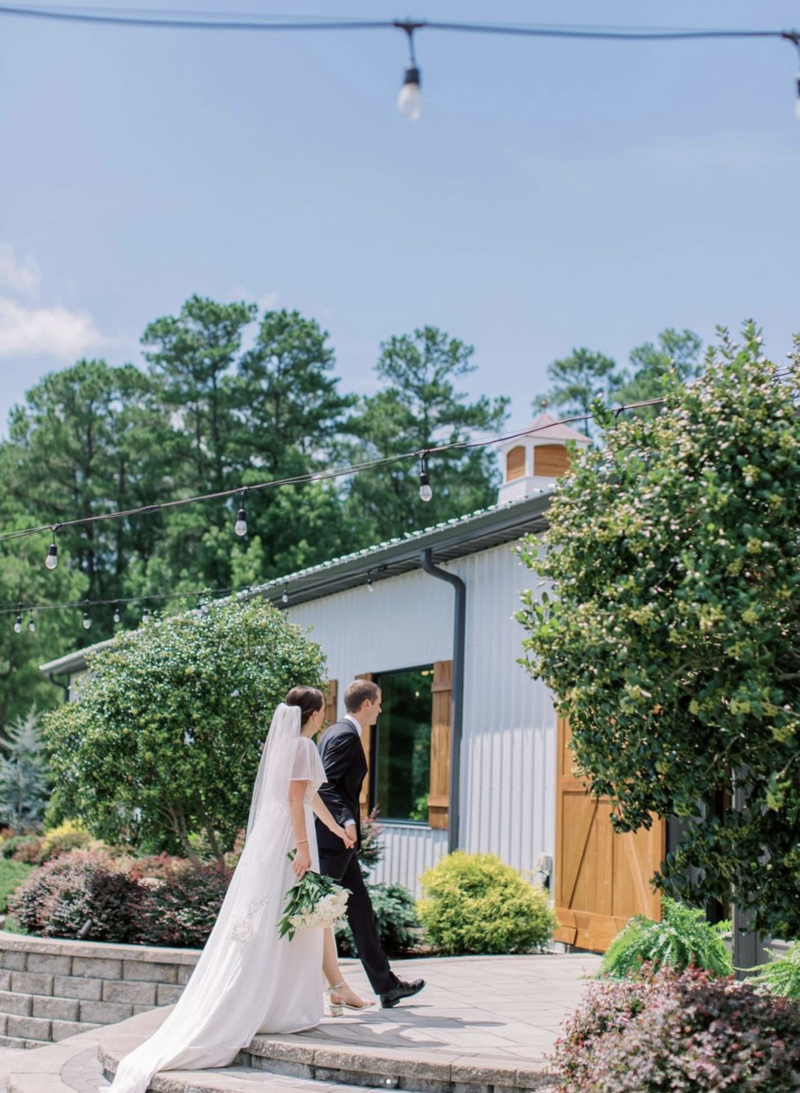 A bride and groom walk hand in hand toward a charming outdoor wedding venue, surrounded by greenery and peaceful garden elements, preparing for their wedding celebration.