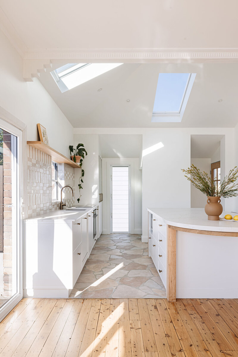 Bright kitchen with skylights, white cabinets, and a stylish island topped with a vase on the wooden floor. The doorway leads outside, embodying the elegance of Luxury Home Design Newcastle.