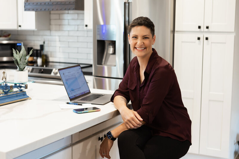 Woman in burgundy blouse sits at kitchen island smiling while working on her laptop