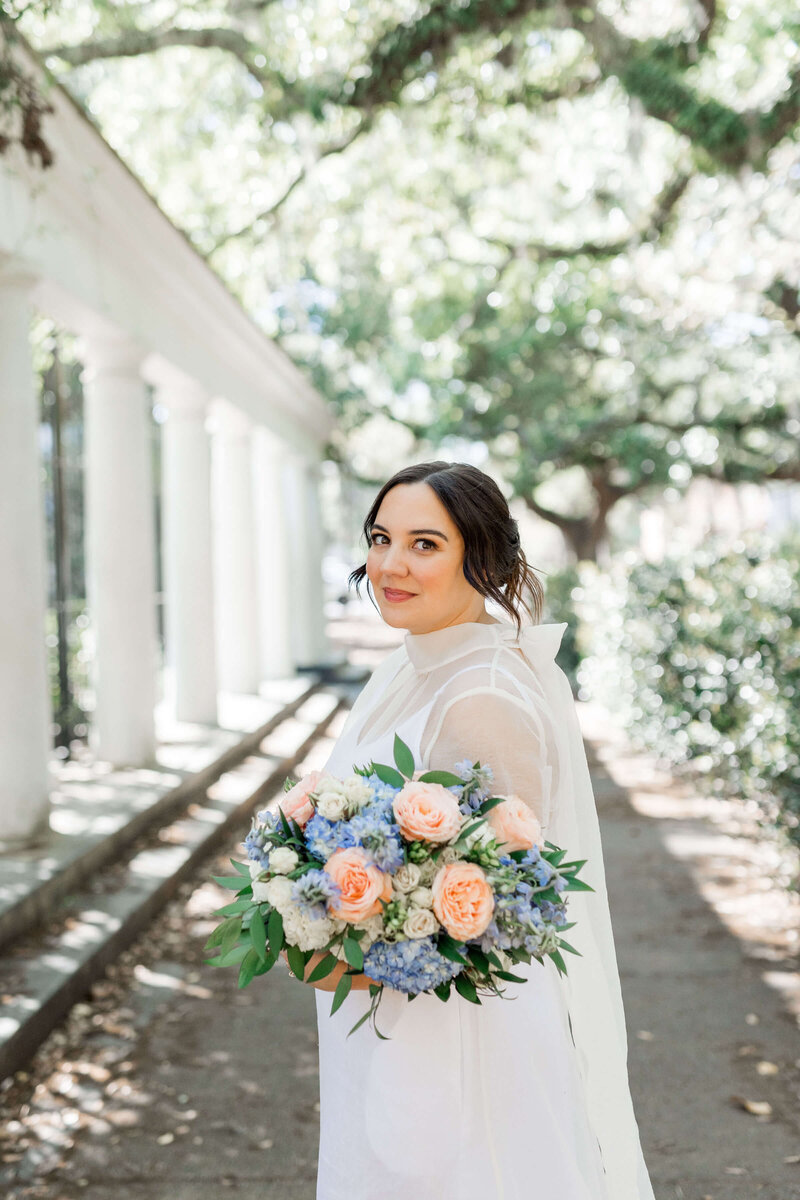 Bride holding a pastel bouquet with peach and blue flowers, standing near white columns and greenery in Savannah, GA, during her elopement.