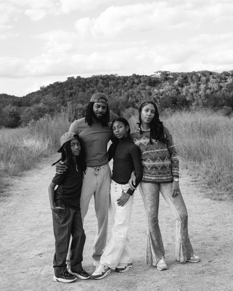 A black and white family portrait of me, my husband, our daughter, and our son standing together on a natural pathway surrounded by grass, distant trees, and clouds. Captured by photographer Maddie Nicole, the image reflects connection and calm in an open outdoor setting.