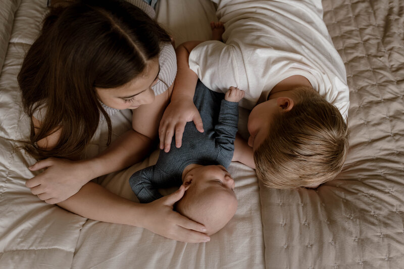 siblings laying with newborn on a bed