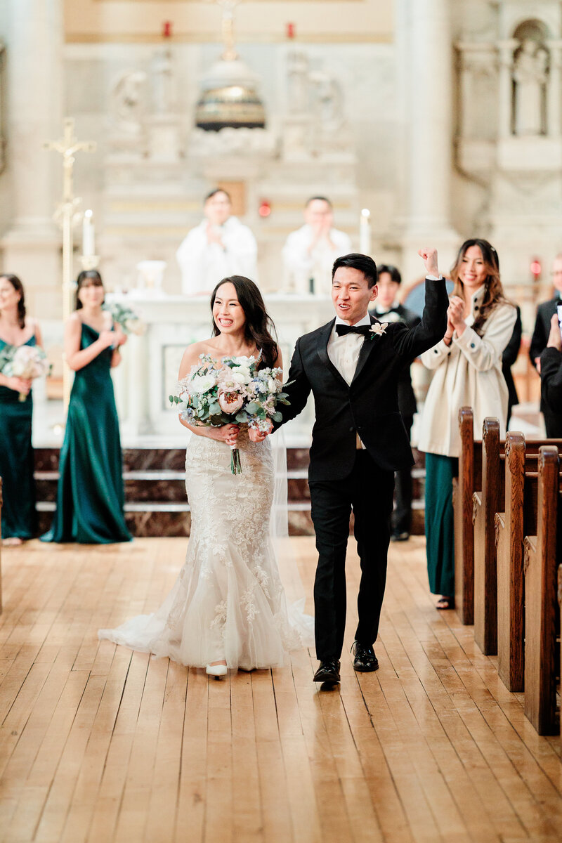 Groom throws fist in the air in celebration while holding the bride's hand as they walk down the aisle after ceremony.