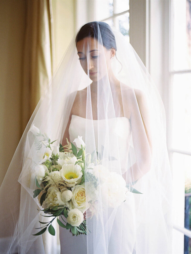 Bride under her veil looking at her flowers as she stands by a window.