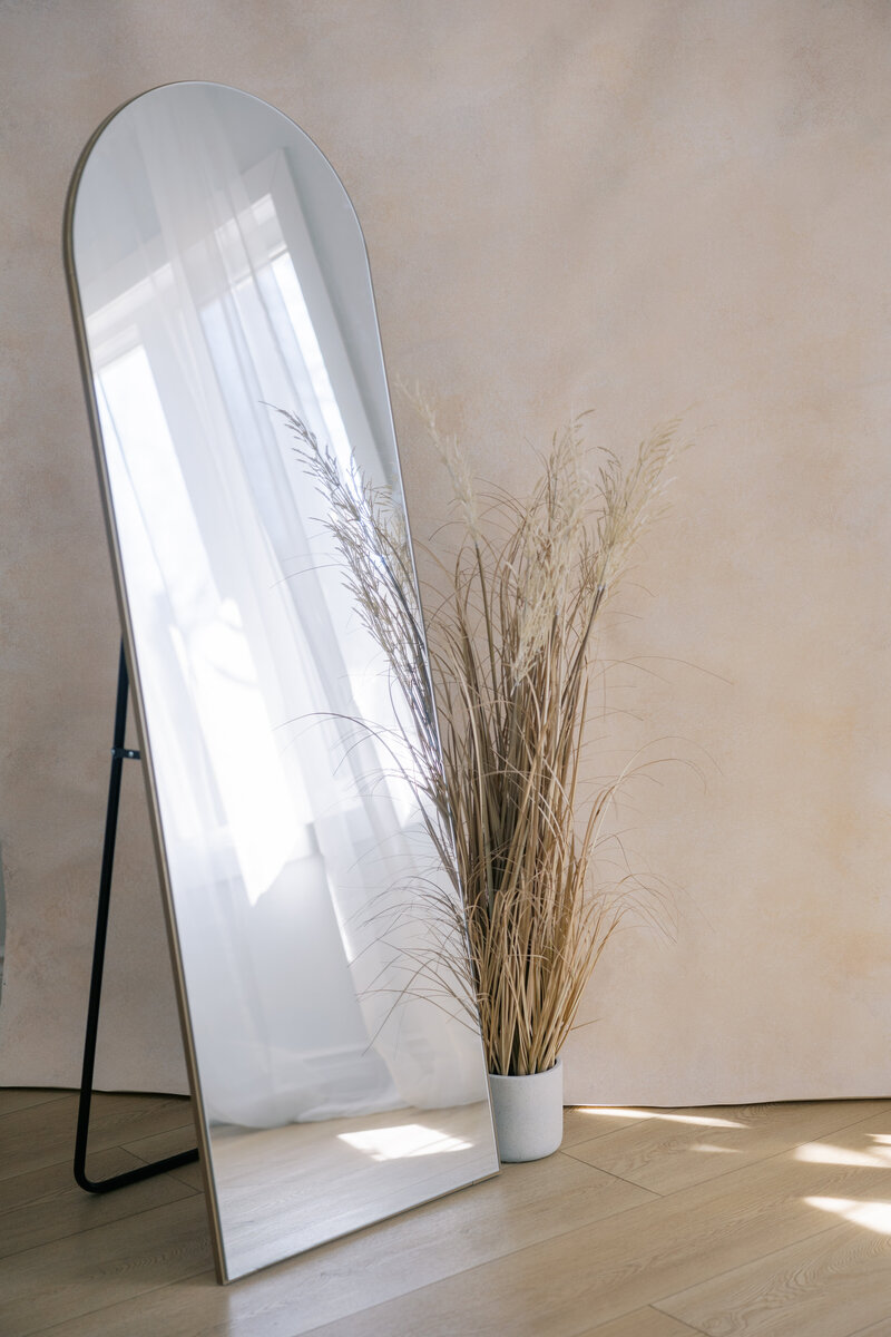 A creamy handpainted background laid on the light wooden floors with a floor length mirror standing up and a white potted plant with taupe grass in it