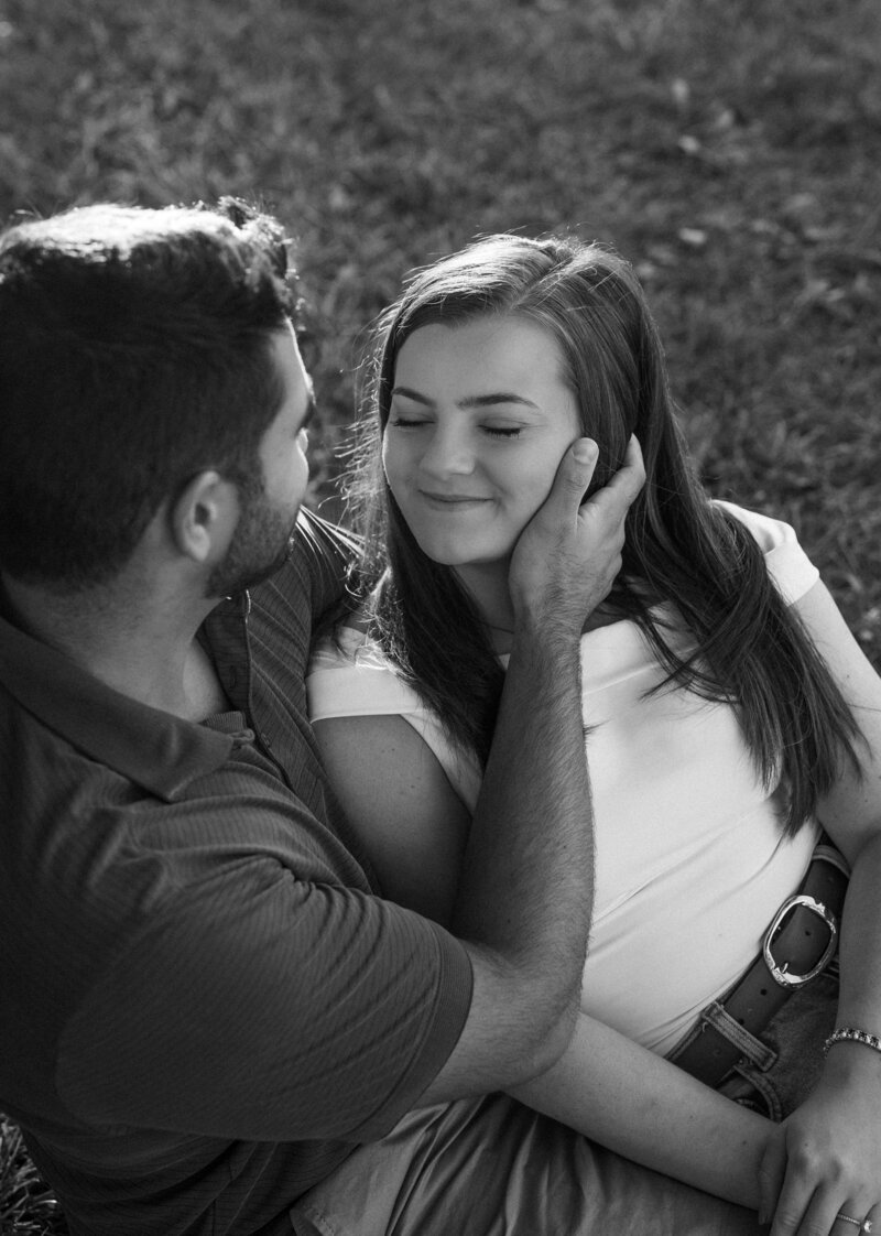Black and white photo of an engaged couple sitting close in the grass at Coxhall Gardens, Carmel