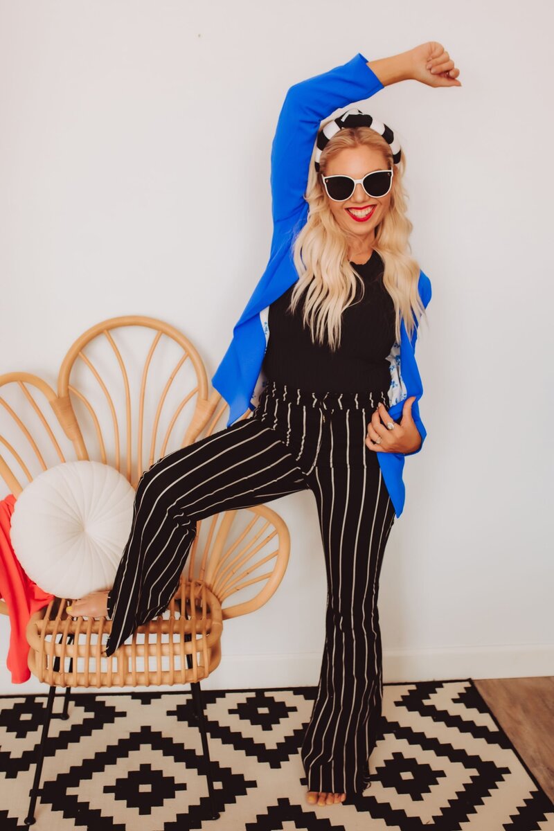 Andi Sweeny of Infitinite Productions wearing a blue blazer, black shirt, and black and white pin stripe pants smiling at the camera with her arm in the air and one leg raised on a chair.