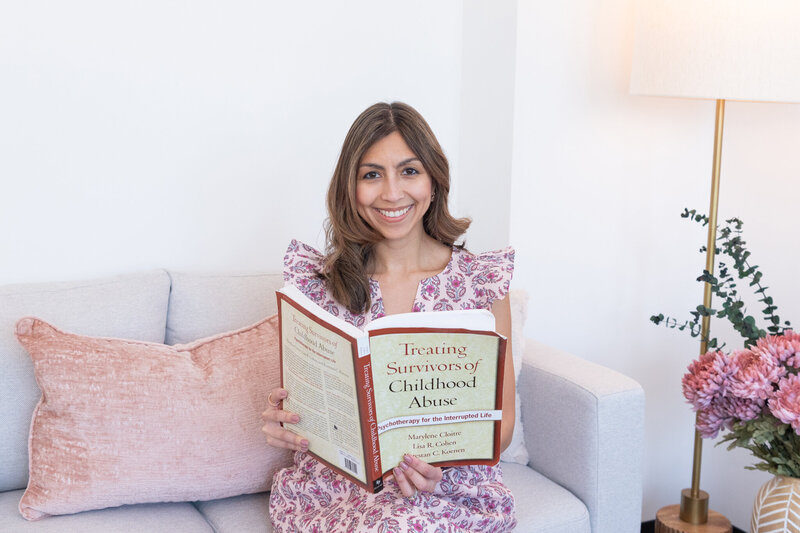 A New Jersey female psychologist specialize in trauma, anxiety, & depression  is sitting on a couch and holding a book about treating survivors of childhood abuse.
