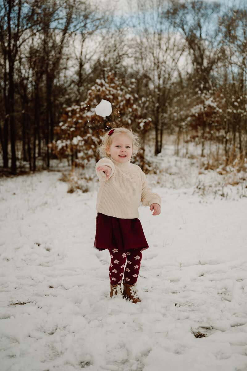 3 year old girl throwing a snow ball towards camera in the snow while wearing burgundy skirt and red sweater in Annapolis Maryland.